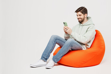 Full body young satisfied happy fun cheerful man wear mint hoody sit in bag chair hold in hand use...