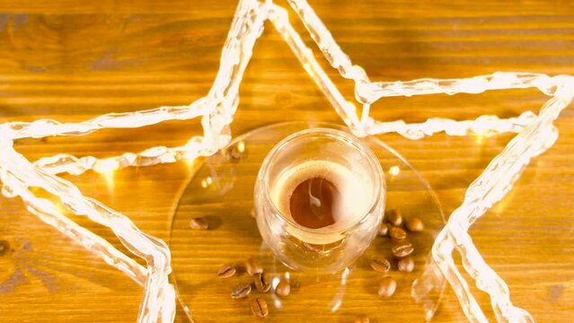 A shot of espresso in a star with Christmas lights and coffee beans around. Steam rises from the coffee. Wooden background.