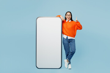 Full body young woman of Asian ethnicity wear orange sweater glasses big huge blank screen mobile cell phone smartphone with area point finger up isolated on plain pastel light blue cyan background.