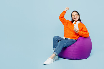 Full body young woman of Asian ethnicity wear orange sweater glasses sit in bag chair hold in hand...