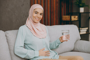 Young muslim woman wears hijab casual clothes drink water hold belly sit on sofa couch stay at home...