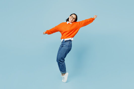Full body side view fun young woman of Asian ethnicity wear orange sweater glasses stand on toes leaning back with outstretched hands dance isolated on plain pastel light blue cyan background studio.