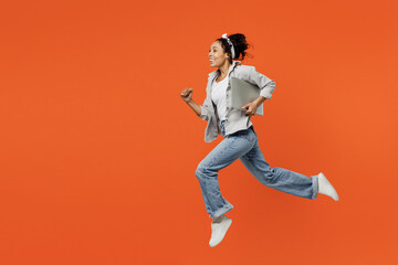 Fototapeta na wymiar Full body fun side view young IT woman of African American ethnicity she wears grey shirt headband jump high holdclosed laptop pc computer run fast hurrying isolated on plain orange background studio.