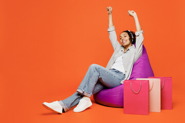 Full body young woman in casual clothes sit in bag chair near paper package bags after shopping raise up hands do winner gesture isolated on plain orange background Black Friday sale buy day concept