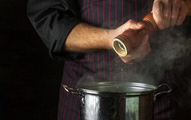 The cook adds ground pepper to a pot of boiling food. Retsoran kitchen cooking concept with advertising space on black background