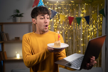 Virtual celebration birthday. Happy guy in party hat celebrating birthday online, blow candle on cake, using laptop for video call with friends or family on webcam at home - Powered by Adobe