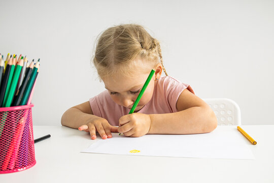 The child girl draws with colored pencils sitting at a white table on a white background