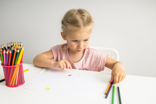 Child girl draws with colored pencils on paper while sitting at a white table