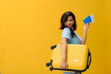 Traveler woman with yellow suitcase, passport and ticket in hand, paper plane, blue t-shirt and jeans on yellow background tourist, travel happiness, glasses, copy space