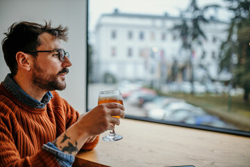 Fototapeta premium Shot of a young tired man drinking beer, using the phone, and looking thoughtfully out of a window.