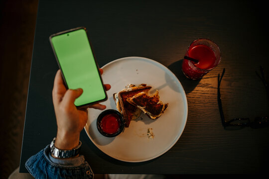 Top view of a male's hands about to eat a sandwich on the plate. Man holding a phone with a green screen on it. - Powered by Adobe