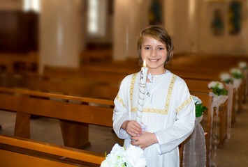 a handsome smiling boy in white clothes with a church candle stands in a Catholic church after...