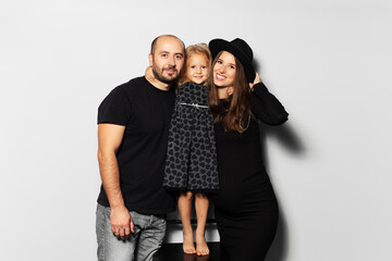 Studio portrait of young happy family, on white background. Wearing hat, dressed in black. Daughter hugging her parents.