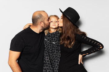 Studio portrait of young happy family, father and pregnant mother kisses their daughter on white background. Wearing hat, dressed in black.