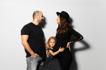 Studio portrait of young happy family, mother and father looking each ever while daughter hugging the belly, on white background. Wearing hat, dressed in black.