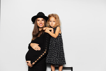 Fototapeta na wymiar Studio portrait of young smiling pregnant woman with her daughter on white background. Wearing hat, dressed in black.