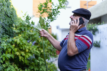 Obraz premium Senior old man talking on the mobile phone while watering his garden