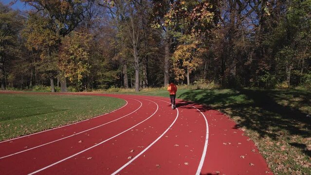 Man in orange sportswear running on race track, aerial drone view. Male runner jogging at stadium. Active sport exercises at autumn day outdoors. Healthy lifestyle
