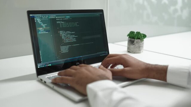 Over-the-shoulder view of smart focused efficient man typing on keyboard using laptop coding. Professional male developer programming apps doing project tasks sitting in bright modern office.