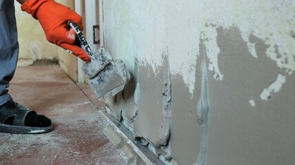 leveling a room wall with a thick layer of plastic putty using a spatula in an orange gloved hand, applying cement mortar on the surface of the wall with a trowel, construction plaster texture