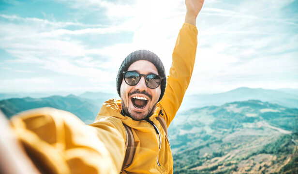 Happy man with backpack taking selfie picture on the top of a mountain - Cheerful hiker climbing cliff - Travel blogger influencer recording adventure live stream on social media platform