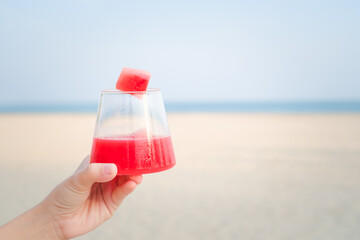 hand holding a glass of watermelon smoothie cocktail by the sea with blue sky background on tropical beach.