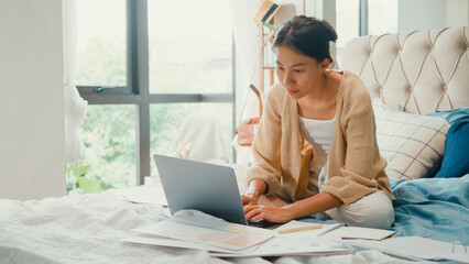 Happy young Asian girl with white cream cardigan on bed focus computer laptop full of paperwork messy document work idea in cozy bedroom at home in morning. Stay quarantine, Work from home concept.