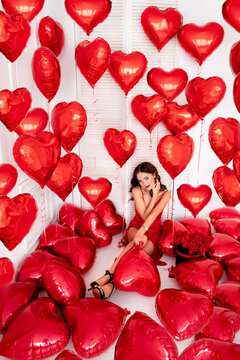 young beautiful woman is sitting surrounded by red balloons. A brunette girl in a red dress. Valentine's Day Concept