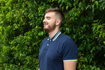 Portrait of a beautiful young bearded man with beard standing on an Ivy wall or in greenery, smiling against from plant, guy relaxing outdoors at sunny summer day. Love nature, eco life