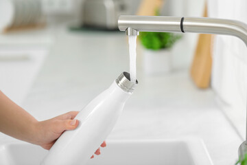 Woman pouring fresh water from tap into thermo bottle in kitchen, closeup