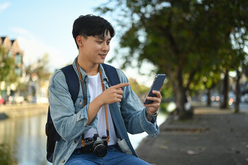 Asian man traveller using smart phone while sitting in the city park on beautiful day. Travel, vacation and journey concept