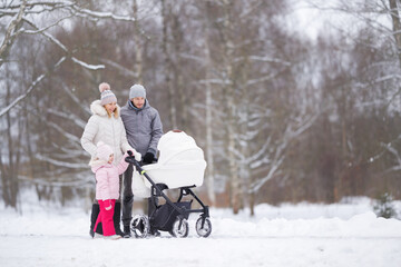 Fototapeta na wymiar Young adult parents and little daughter pushing white baby stroller and walking on snow covered sidewalk at park in cold winter day. Spending time together. Two child family.