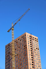 Fototapeta premium Tower crane and unfinished building on background of blue sky, vertical shot. Housing construction, apartment blocks in city