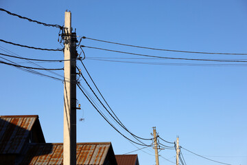 Powerline posts with electrical wires and capacitors above old roofs on blue sky background....