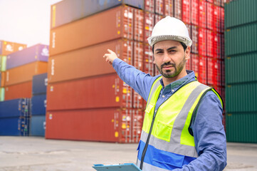 Portrait of handsome caucasian man with white helmet and reflection shirt, holding document paper and pointing finger in front of container cargo store. import and export logistic industry concept.