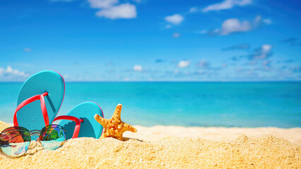 Beautiful colorful background for summer beach holiday. Sunglasses, starfish, turquoise flip-flops on sandy tropical beach against blue sky with clouds on bright sunny day.