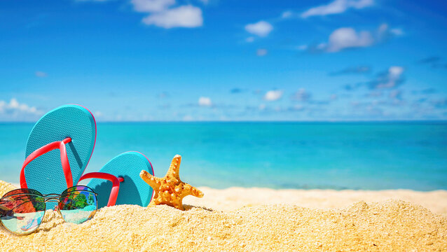 Beautiful colorful background for summer beach holiday. Sunglasses, starfish, turquoise flip-flops on sandy tropical beach against blue sky with clouds on bright sunny day.