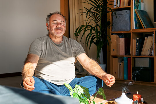 Overweight man practicing yoga and meditation at home sitting in lotus pose on yoga mat, relaxed with closed eyes. Mindful meditation concept. Wellbeing.