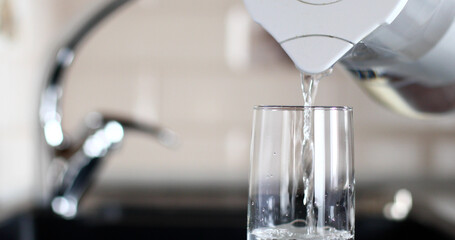 Pouring water from the filter into a glass standing on the table next to the sink, with a kitchen faucet in the background. The concept of using clean water