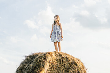 Naklejka na ściany i meble Pretty little girl stand at top of haystack looking away. Beautiful blue sky. Freedom. Summer rest, countryside life.