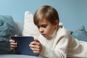 Boy spends time behind a screen on a tablet on the sofa in the living room. Digital device. Technologies.