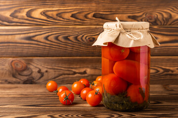 Pickled red cherry tomatoes in a glass jar on brown textured rustic wood. Food preservation for autumn or winter. Home storage. Canning, fermentation concept. Place for text. Copy space.