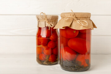 Pickled red cherry tomatoes in a glass jar on a white textured rustic wood. Food preservation for autumn or winter. Home storage. Canning, fermentation concept. Place for text. Copy space.