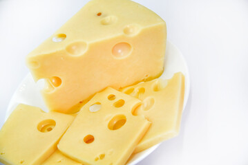 A large piece of cheese with holes on the plate. Chopped cheese and small slices on a white background. Juicy and delicious natural cheese. Cheese holes.