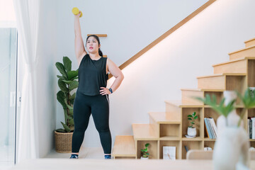 fat woman standing lifting dumbbell workout in living room. chubby woman raising arm using dumbbell...