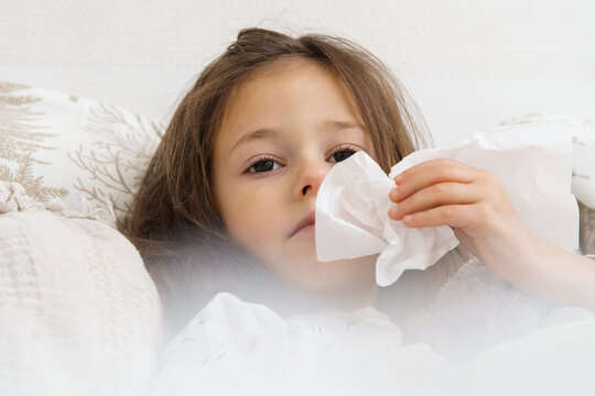 Little girl blow stuffy nose with white handkerchief. Portrait closeup of unhealthy child, selective focus. Kid have bed rest. Home treatment. Runny nose, snot, cold symptoms, cure.