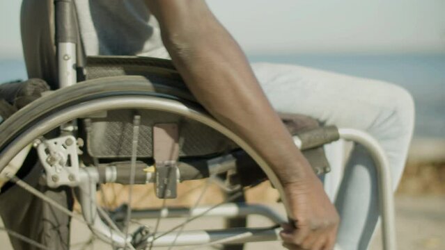 Closeup of Black man with paraplegia riding wheelchair along seafront on sunny day. Strong male arms pushing wheels. Blurred seascape background. Side view. Disability, lifetsyle, motivation concept.