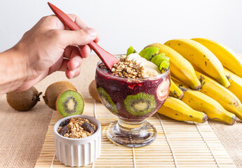 Acai, Brazilian acai bowl with kiwi, banana and oat flakes, hand holding a wooden spoon for eating.