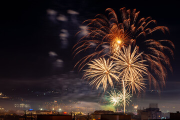 Festive firework bursts in the night sky. Yellow orange fireworks explosions over city celebrating holiday event. Pyrotechnic for celebration concept. Night scene. Long exposure shot.