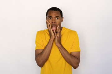 Scared young man holding his face with hands from shock. Male African American model with brown eyes and short black haircut in yellow T-shirt seeing something scary. Fright concept